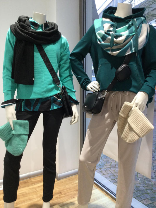 Zwei Schaufensterpuppen in Wintermode: links in Türkis und Schwarz mit Schal und Mütze, rechts in Petrolgrün mit Hoodie, Schal und beiger Hose